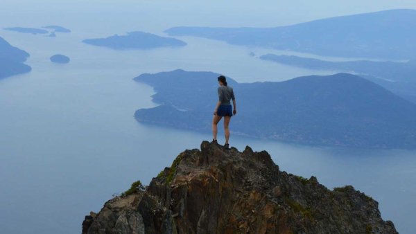 A person stands on top of a mountain overlooking the blue waters of Howe Sound fjord.